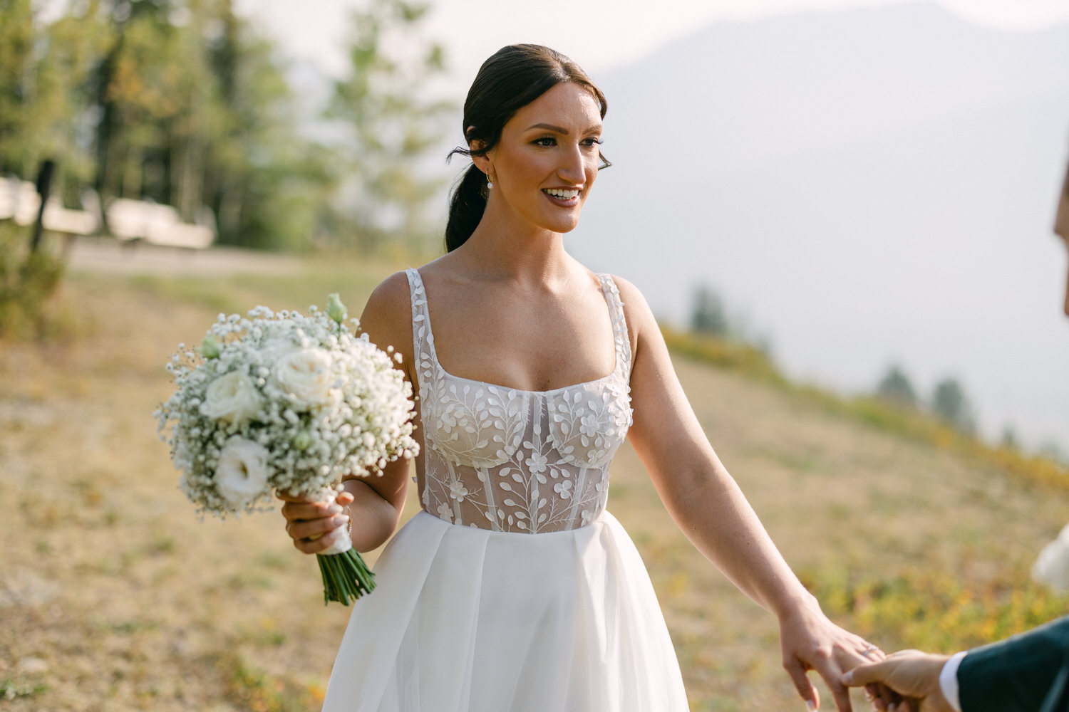 A bride in an intricate gown holding a bouquet, smiling while reaching for her partner against a serene outdoor background.