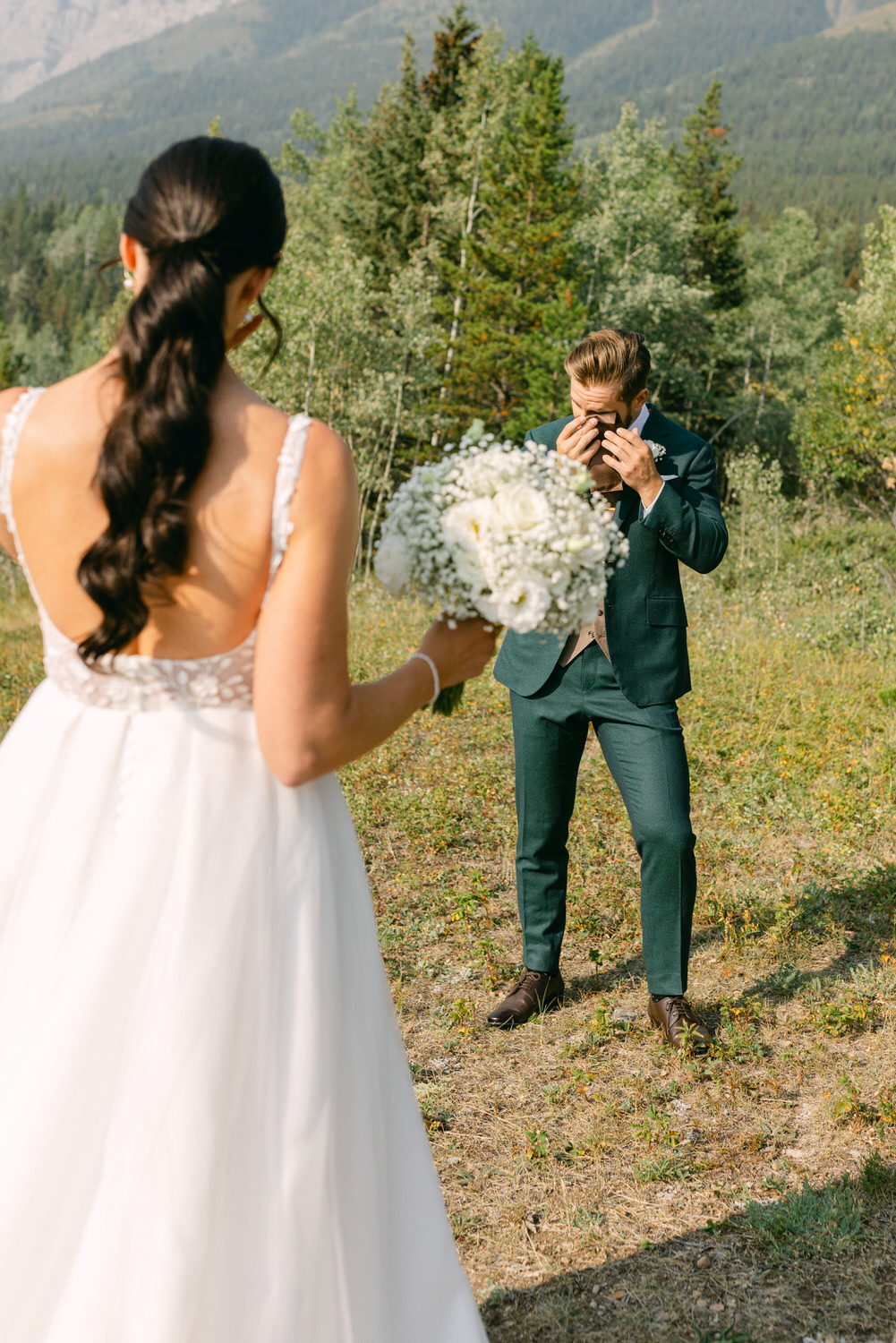 A bride in a stunning dress holds a bouquet while the groom, visibly emotional, wipes his eyes in a beautiful outdoor setting surrounded by trees and mountains.