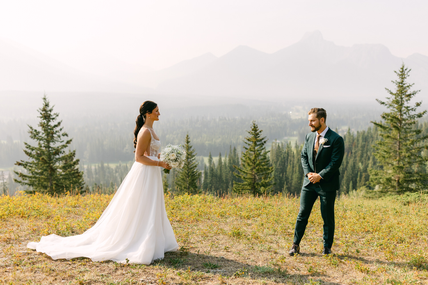 A bride in a flowing white gown holding a bouquet stands before a groom in a dark suit, both surrounded by a breathtaking mountain landscape.