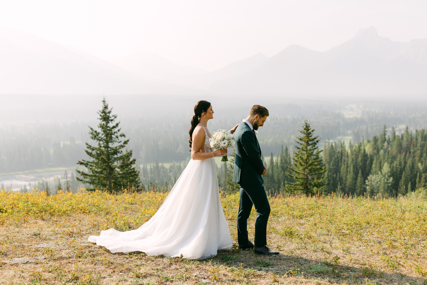 A couple standing in a scenic mountainous backdrop, with the bride in a flowing white dress holding a bouquet, while the groom stands pensively with his hands clasped.