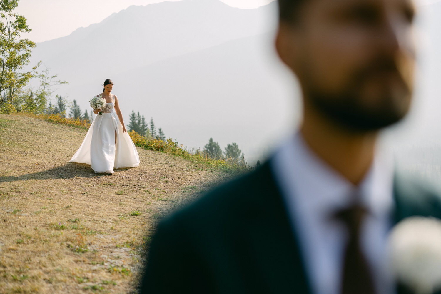 A bride in a flowing white gown walks towards the camera holding a bouquet, set against a backdrop of mountains and soft sunlight. A blurred figure of a man is seen in the foreground.