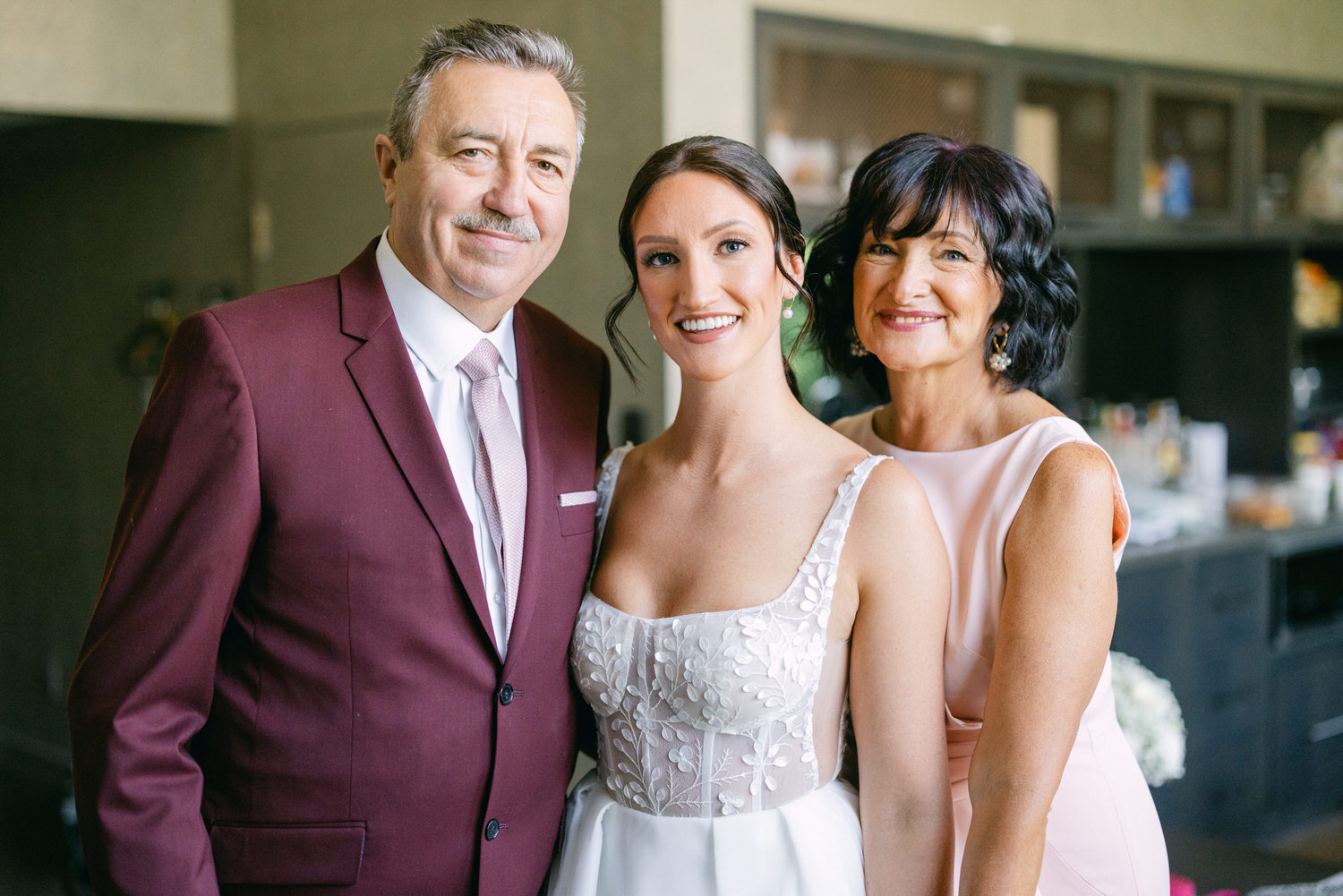 A bride poses with her smiling parents in elegant attire, showcasing a warm family bond in a stylish interior setting.