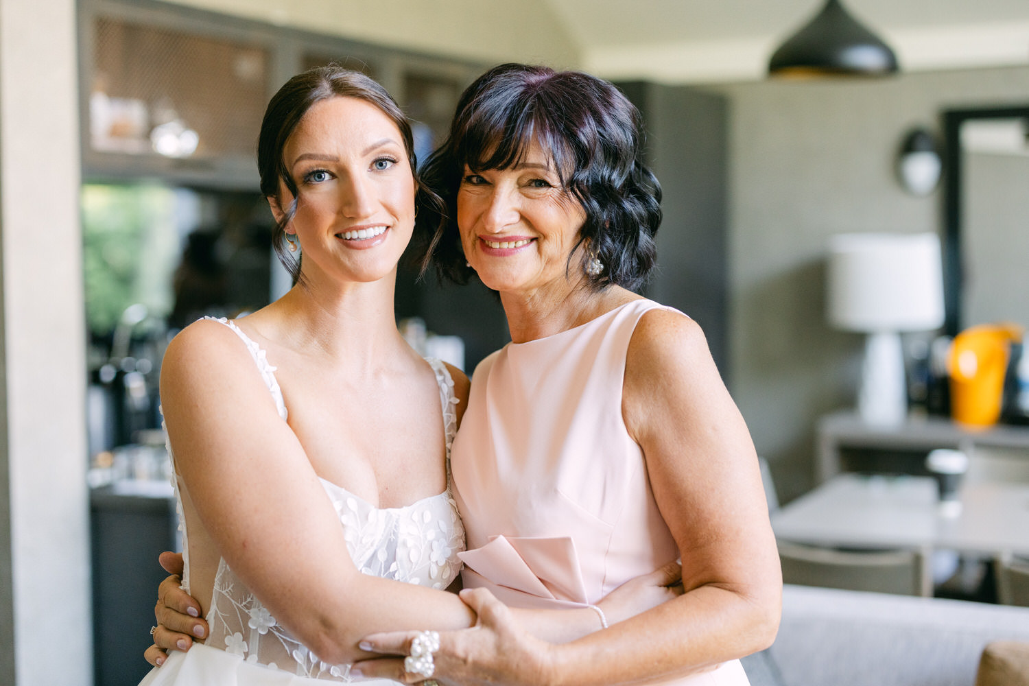 A smiling bride in a floral lace gown poses with her mother, who is wearing a light pink dress, in a well-lit modern room.