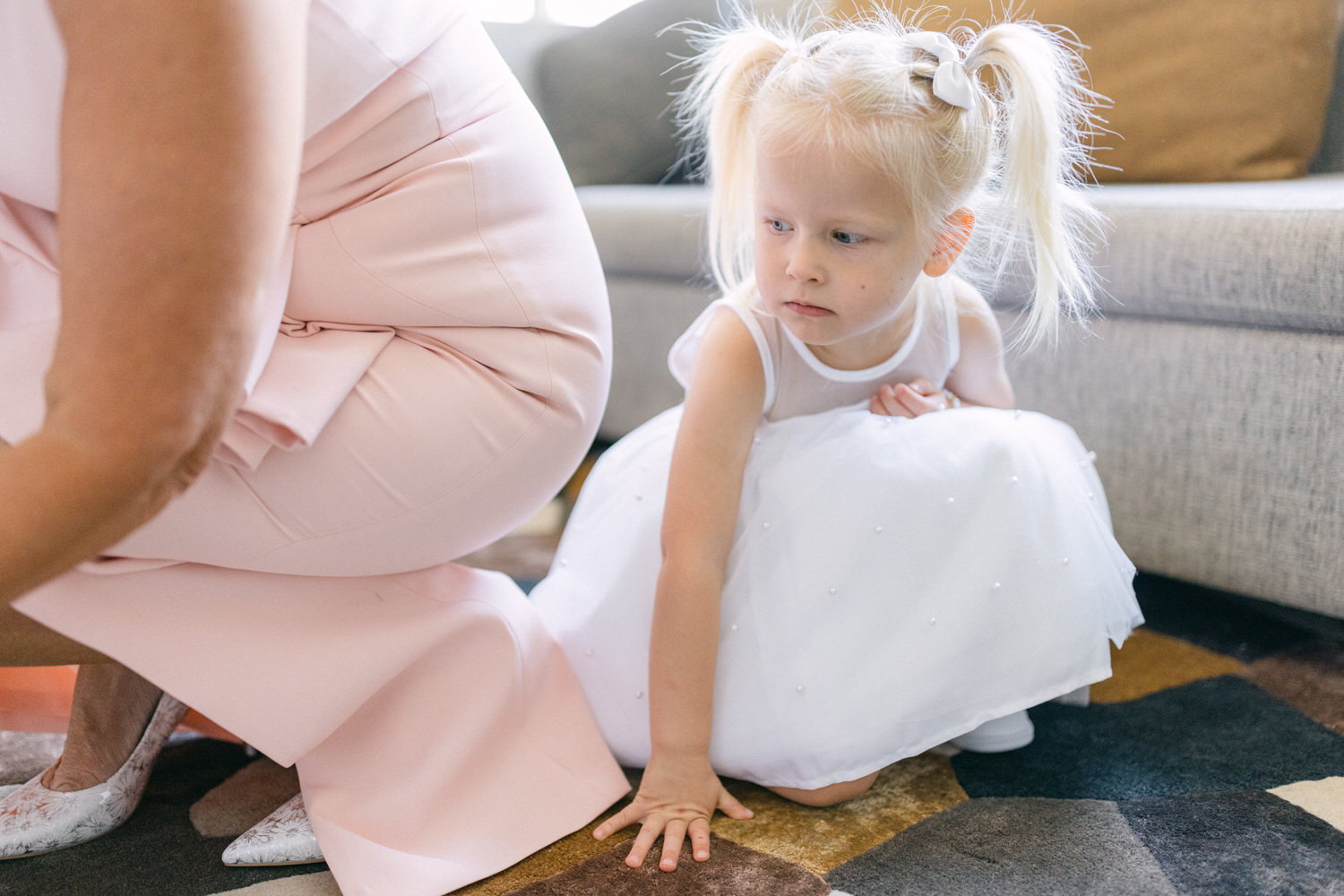 A young girl in a white dress with pearls kneels on the floor, looking thoughtfully as she adjusts her outfit, while an adult in a pink outfit is partially visible in the background.