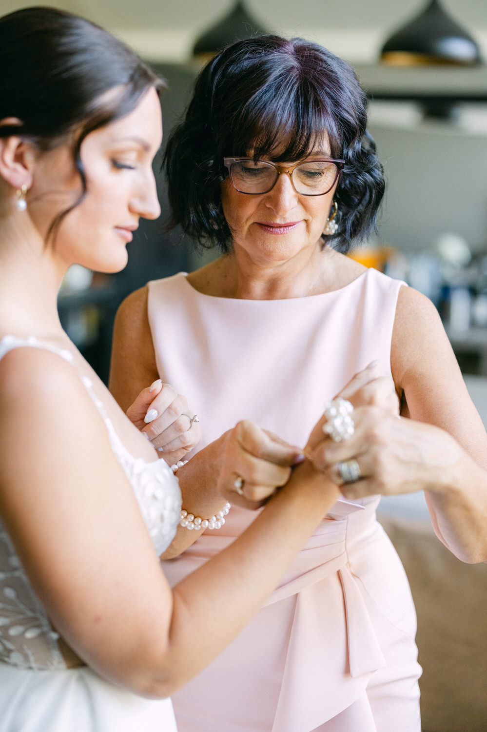 A woman helps another woman with a bracelet, reflecting a tender moment before a special occasion, with soft lighting and elegant attire.