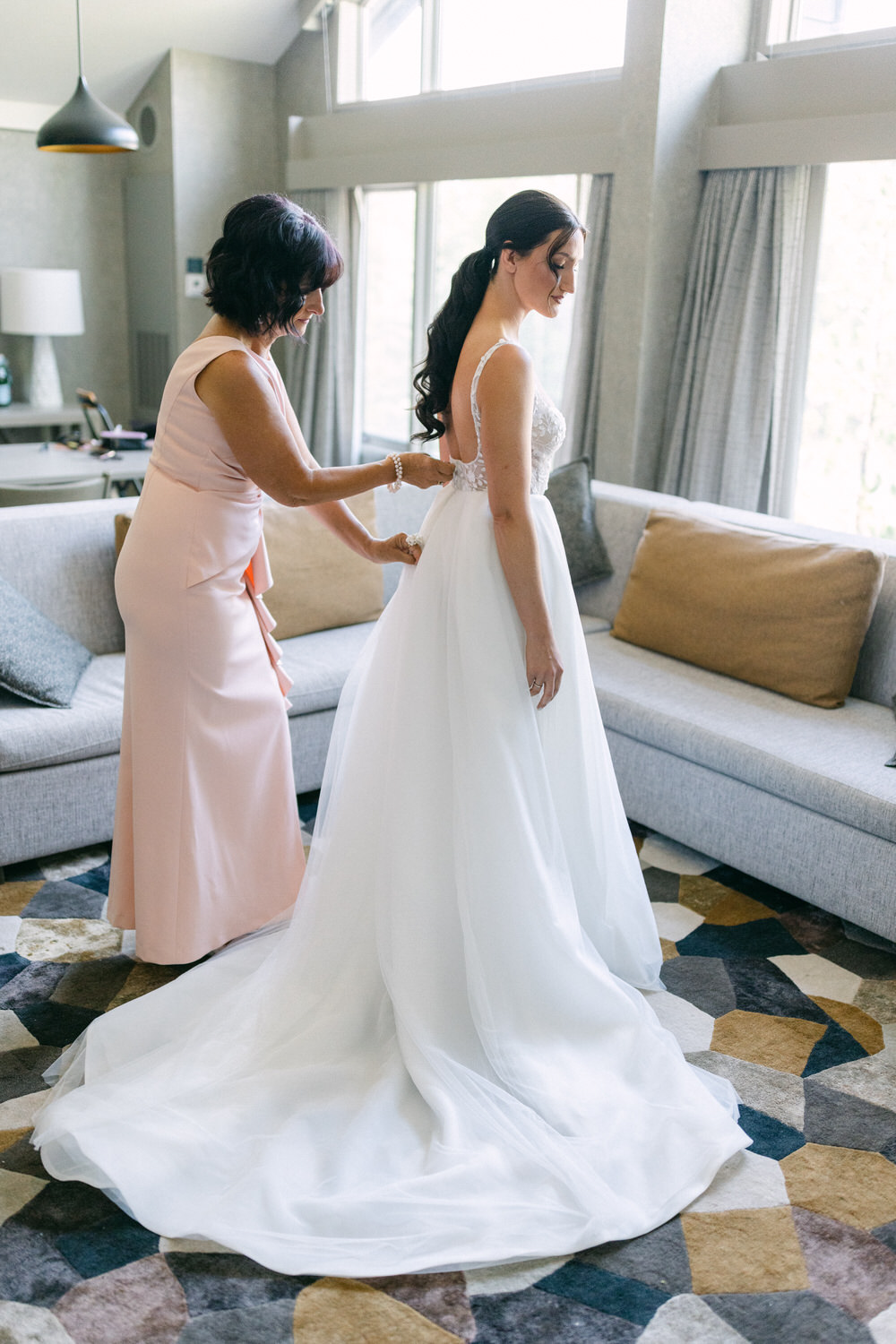 A bride in a white gown is being assisted by a woman in a pink dress as she fastens the back of her dress, surrounded by a stylish, modern living room.