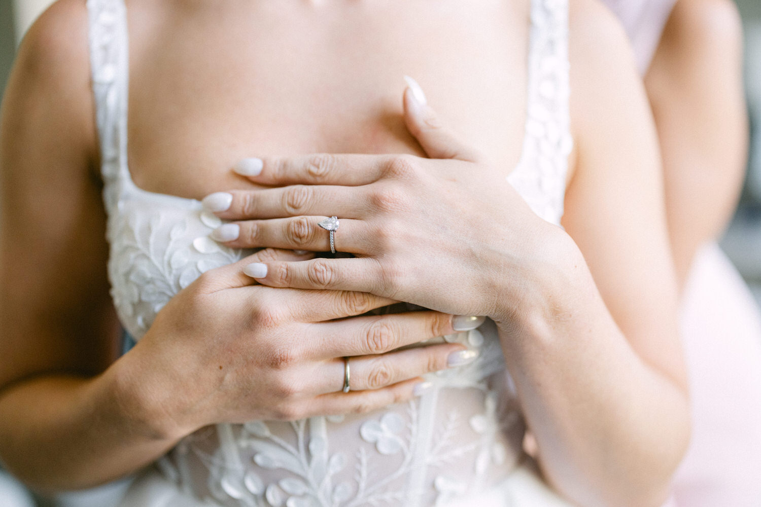 Close-up of a bride's hands showcasing a diamond engagement ring and a wedding band, contrasted against her intricately designed white gown.