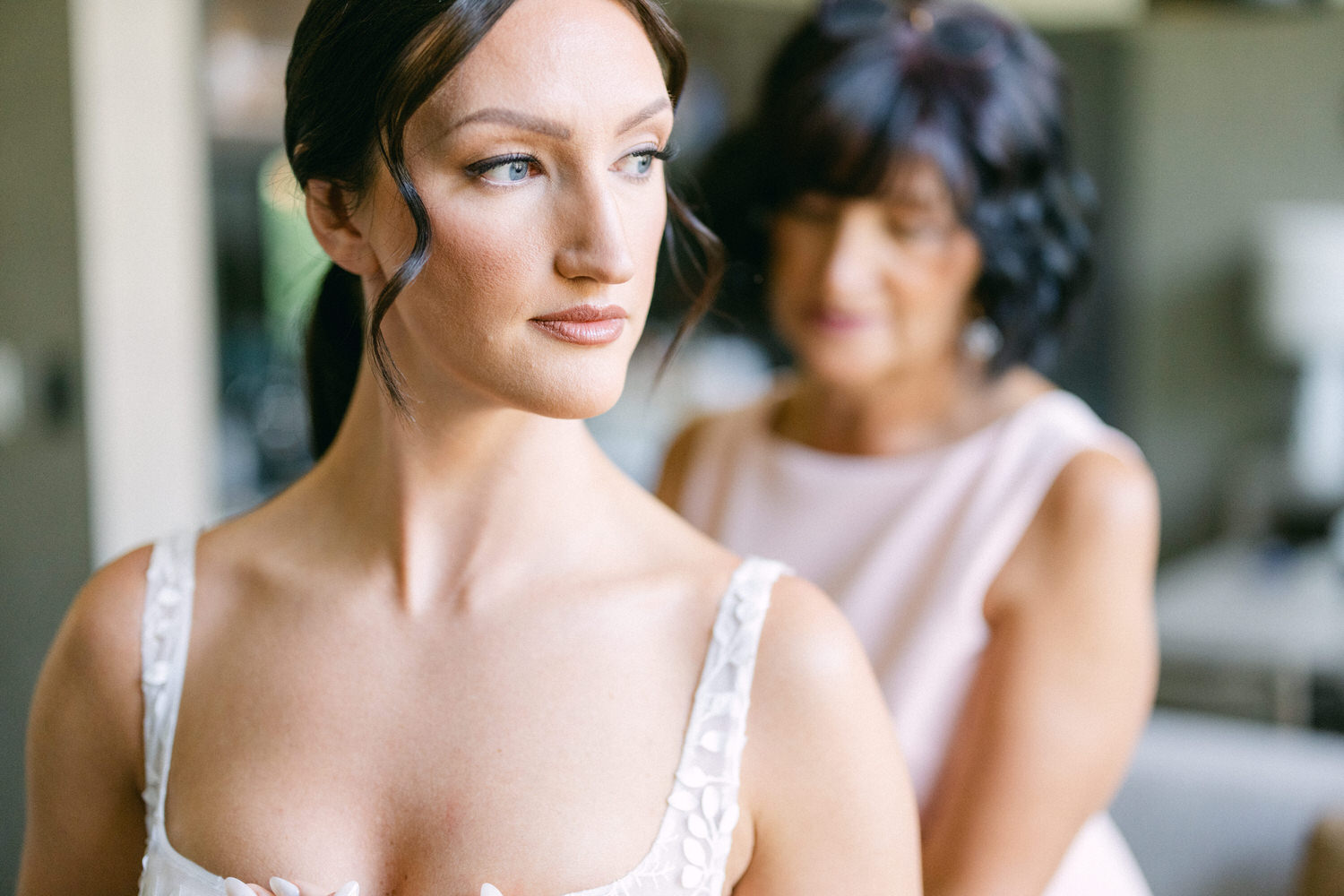 A bride in a delicate lace gown gazes thoughtfully while her mother adjusts her dress in the background.