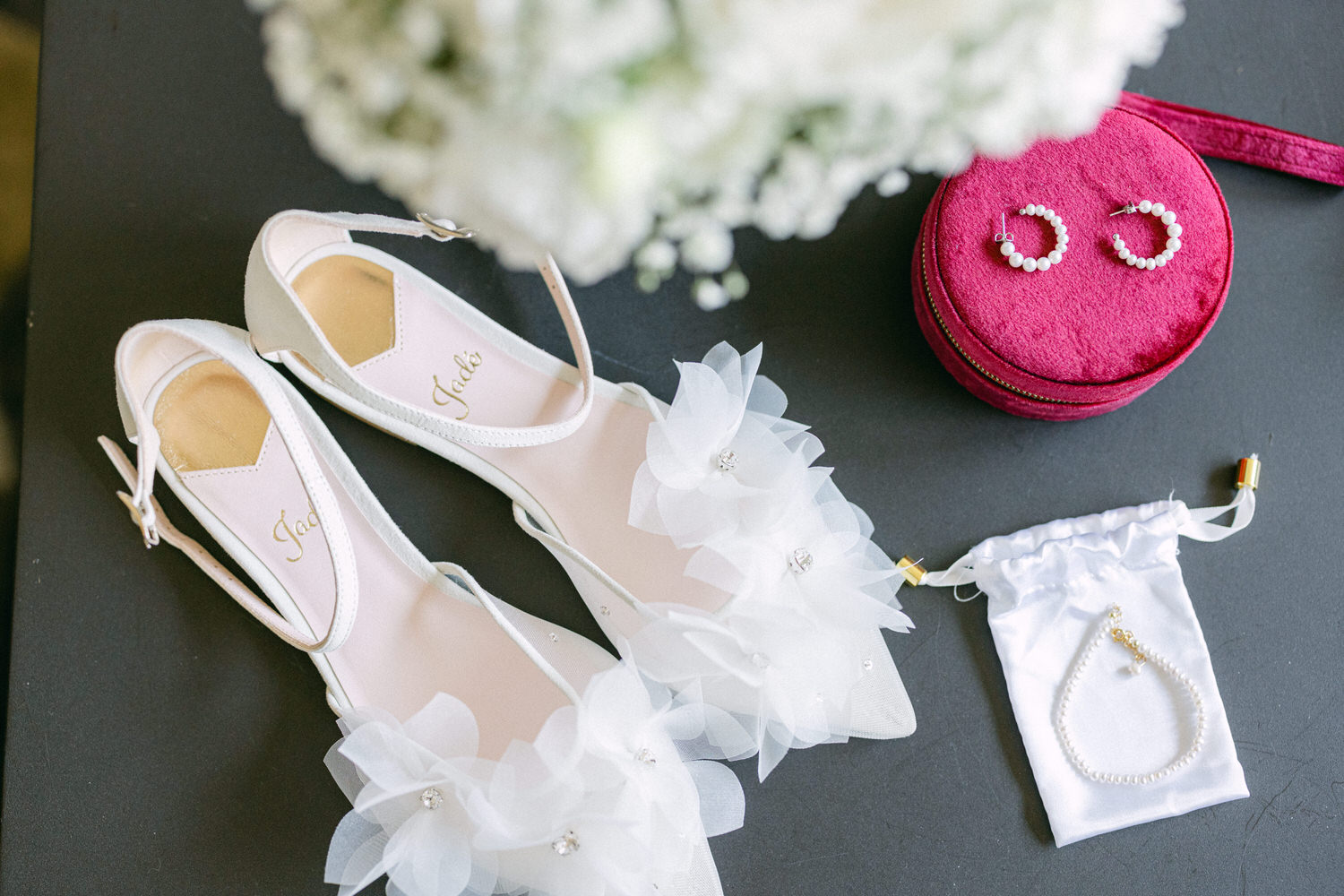 Close-up of white floral wedding shoes, a pink jewelry case with earrings, and a satin pouch containing a bracelet, elegantly displayed on a dark surface.