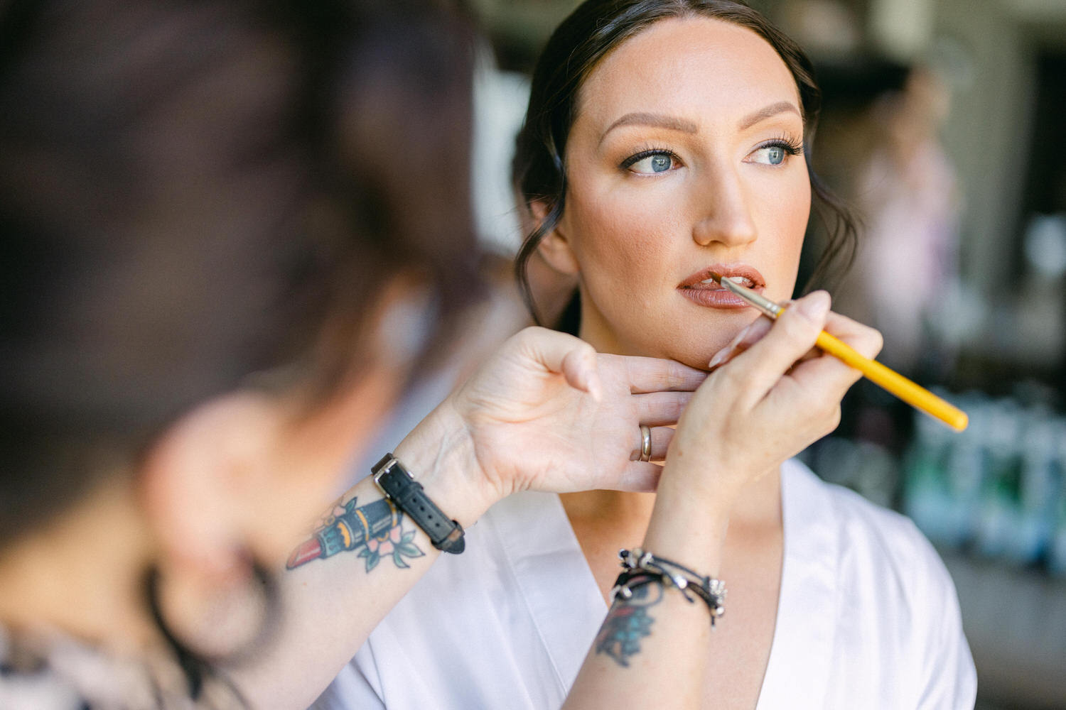 A close-up of a woman having her makeup applied, focusing on the artist's hand using a brush on her lips. The artist's tattoos are visible, adding a personal touch to the scene.