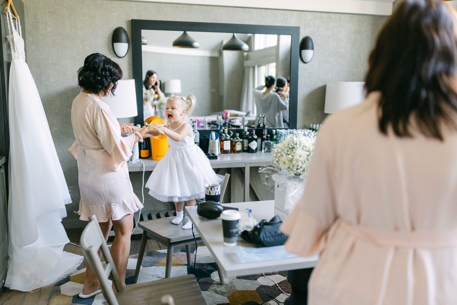 A joyful little girl in a white dress stands on a table, reaching for a woman wearing a robe, as they share a playful moment in a bridal preparation room filled with flowers and beauty products.