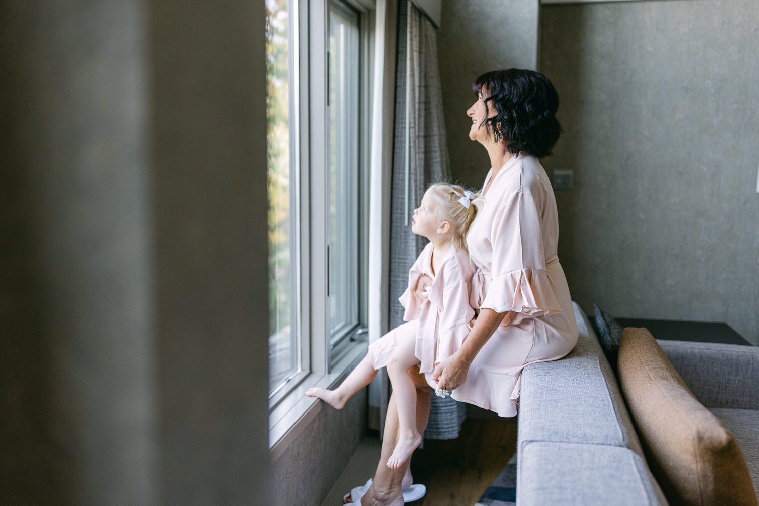 A woman and a young girl in matching pink robes sit by a window, looking outside with joy and curiosity.