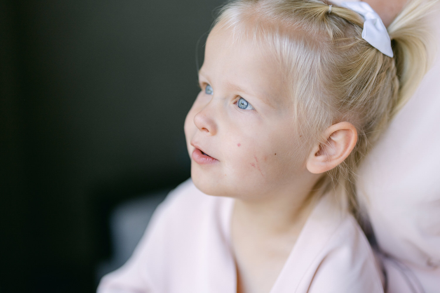 A close-up of a young girl with blonde hair and a white bow, looking thoughtfully to the side while wearing a light pink robe.