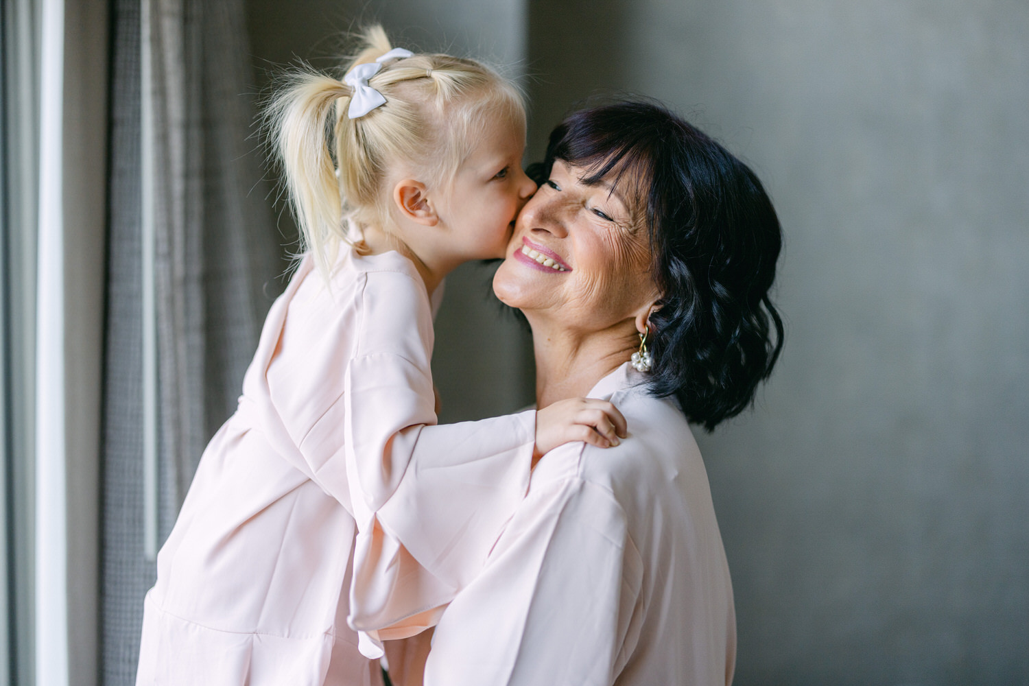 A young girl kisses her grandmother on the cheek, both smiling warmly, radiating love and joy in matching pink outfits.