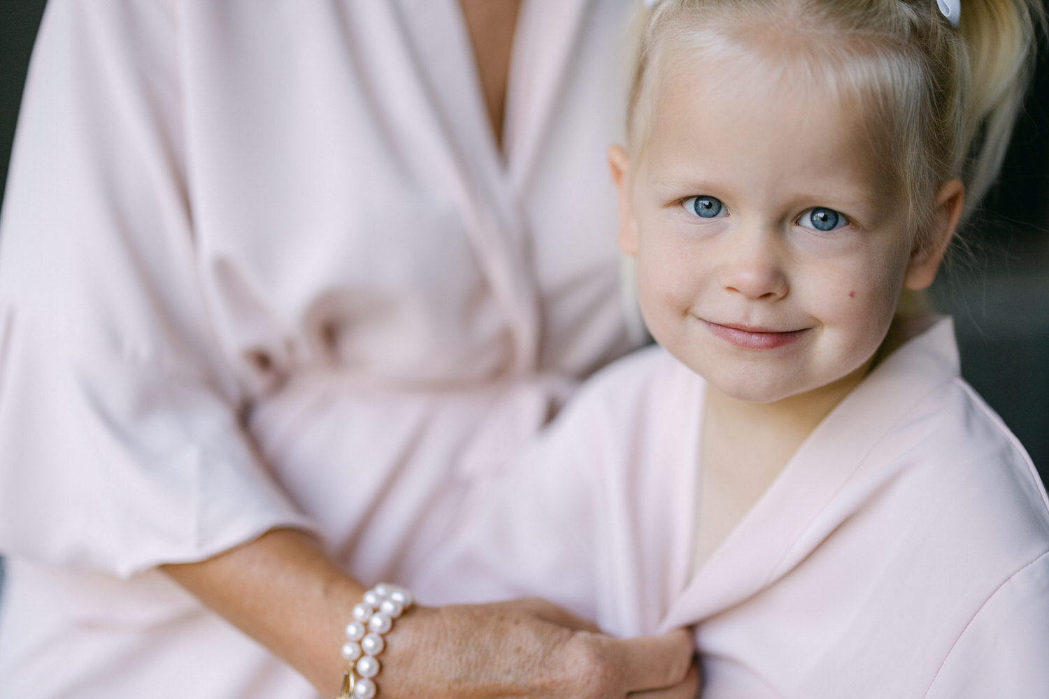 A young girl with blue eyes smiles warmly while being embraced by an adult in a light pink robe.