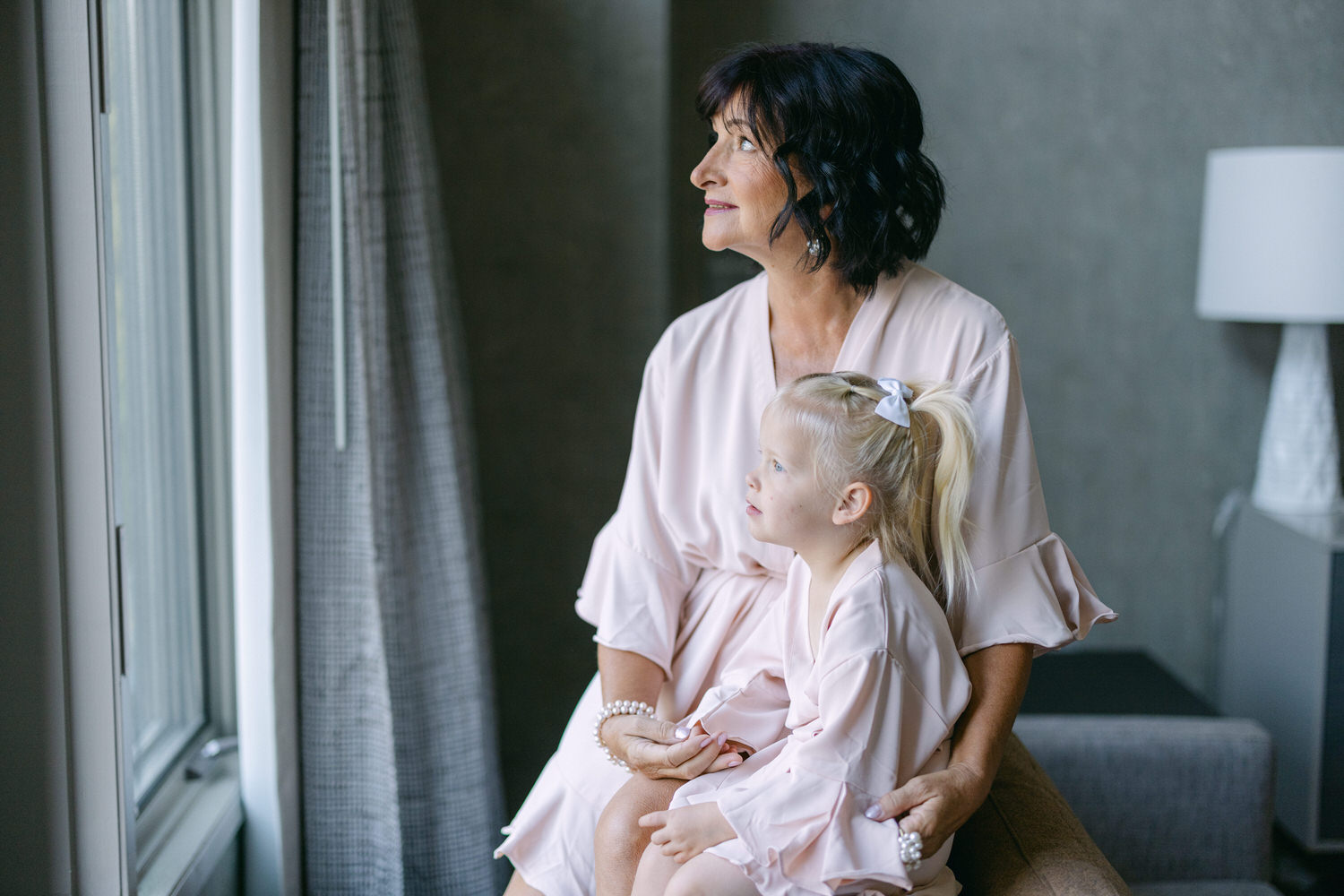 A grandmother and her granddaughter in matching pink robes sit together, gazing out a window, sharing a tender moment of connection and joy.