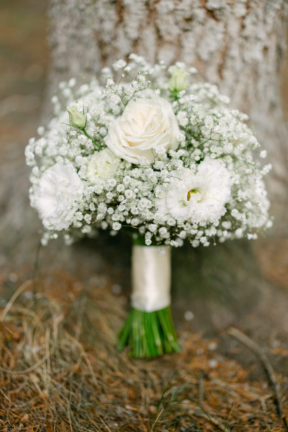 A beautifully arranged bouquet featuring white roses, delicate baby’s breath, and other white flowers, set against a textured tree trunk background.