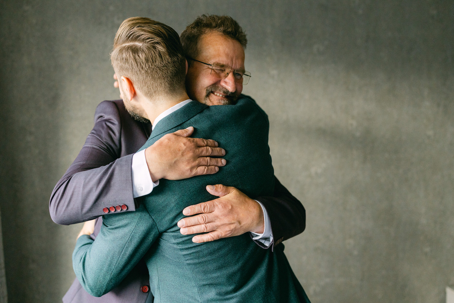 An emotional moment of a father and son sharing a heartfelt hug, both dressed in formal suits against a softly lit backdrop.