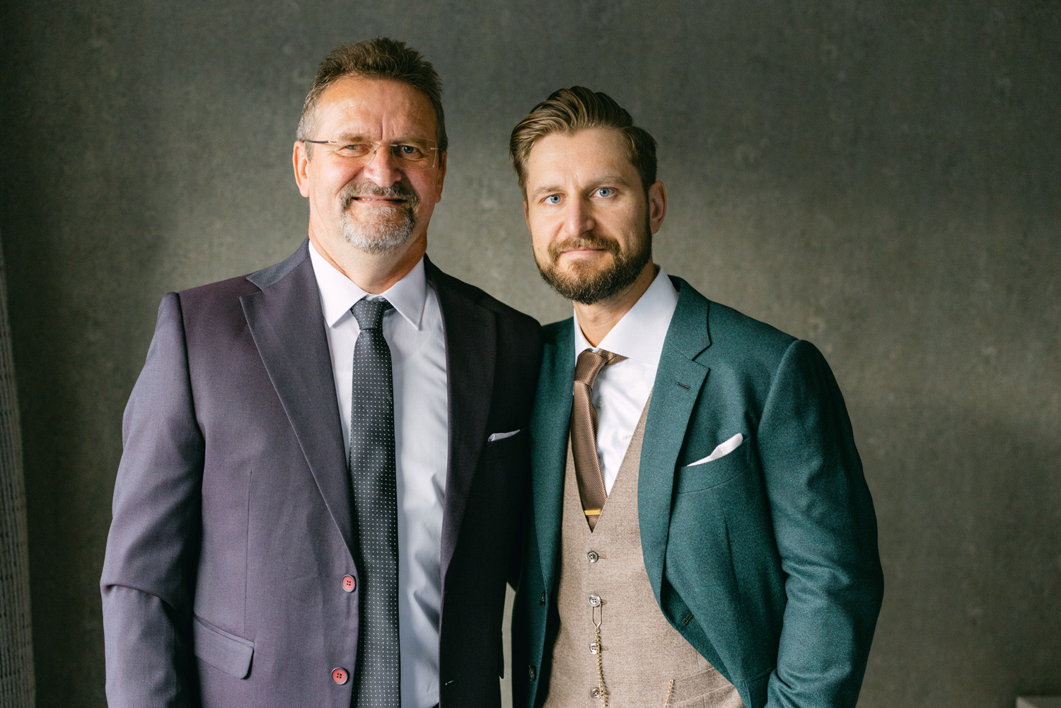 A father and son pose together in formal suits against a neutral backdrop, showcasing their styles and proud expressions.