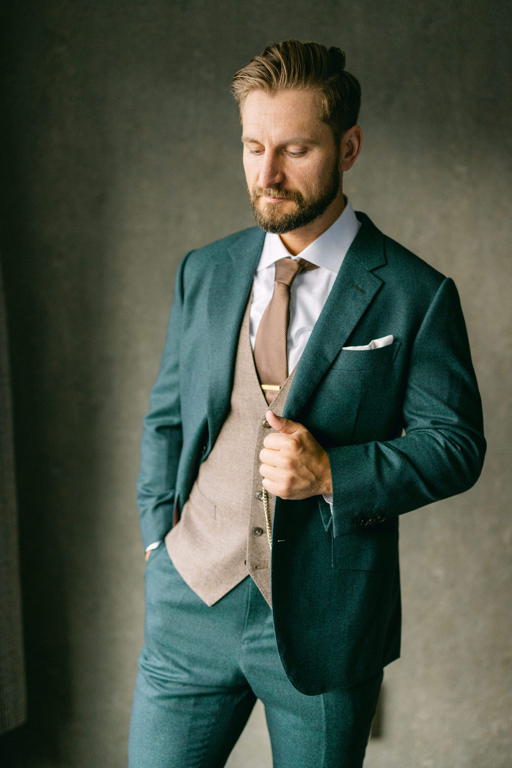 Stylish Man in Suit::A well-dressed man wearing a teal suit with a beige vest and brown tie, presenting a sophisticated look with a neutral background.