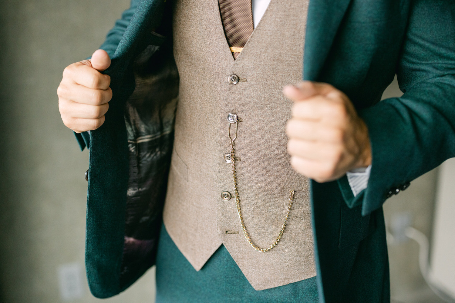 A close-up of a man in a stylish green coat, showcasing a beige vest with a chain accessory, highlighting sophisticated menswear fashion.