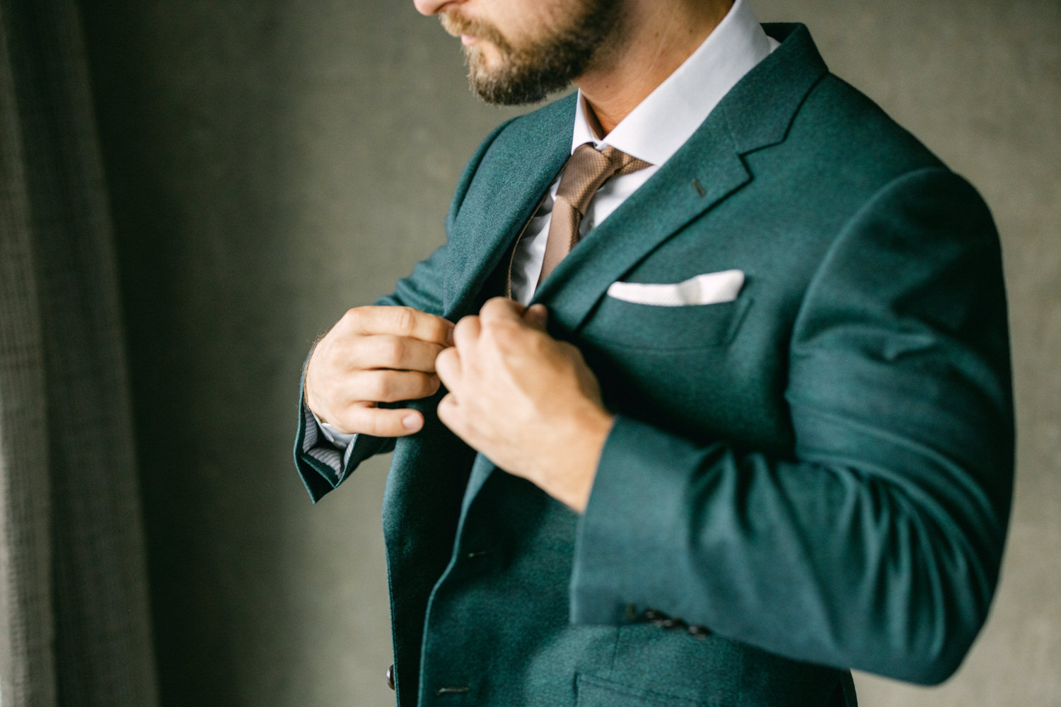 Groom Adjusting Suit::A close-up of a man in a green suit, buttoning his jacket, with a tie and a white pocket square, showcasing a focused expression.