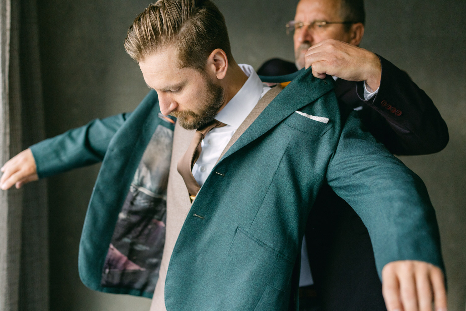 A man dressed in a suit is having a jacket put on by another person in a stylish indoor setting.