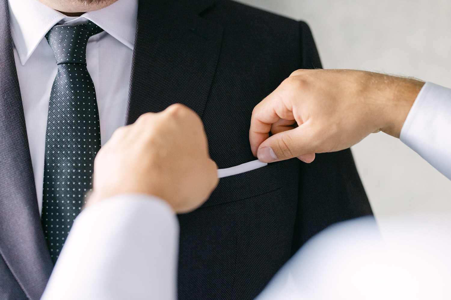 A close-up of a man adjusting the pocket of a dark suit while another person helps with a white accessory.