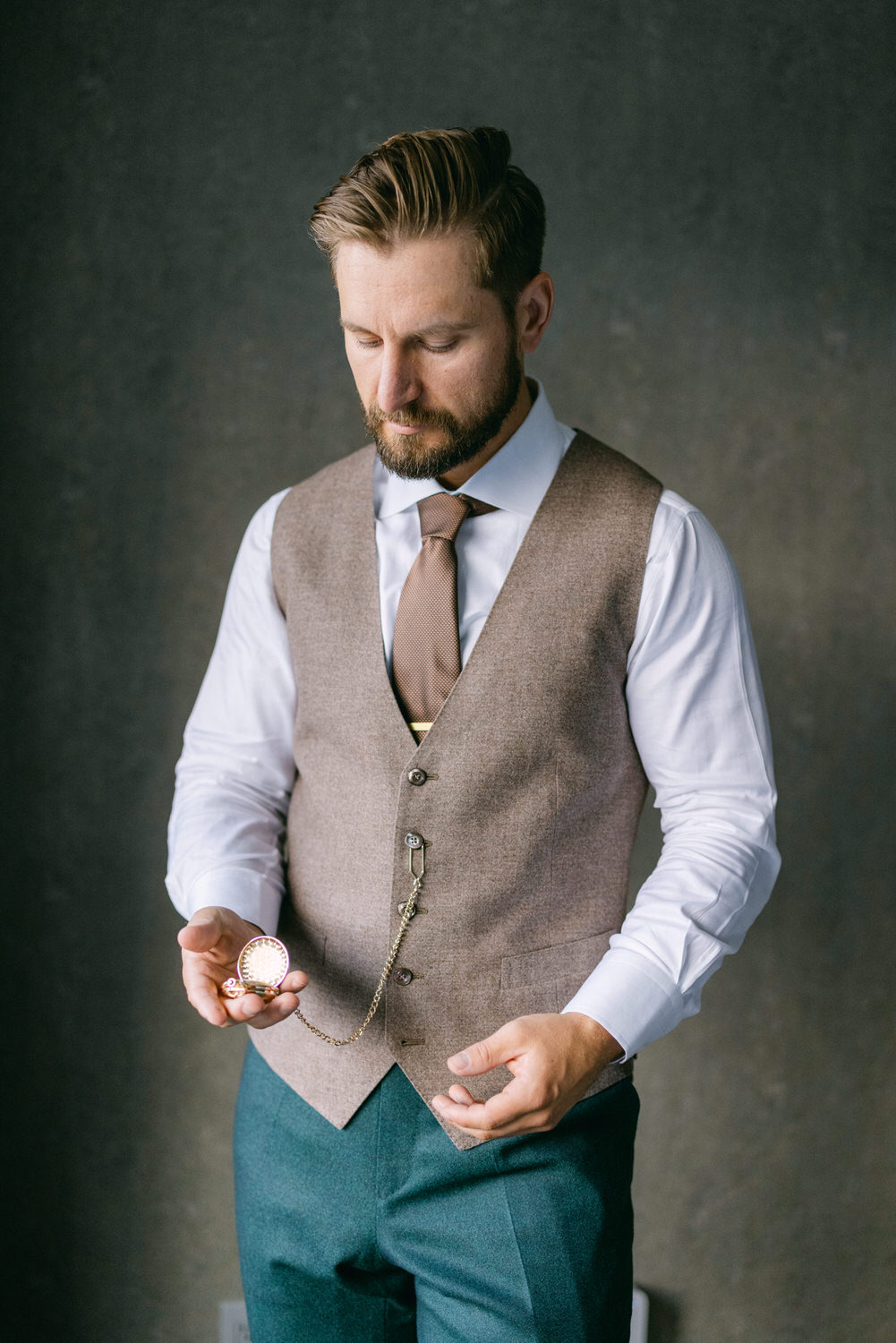 A well-dressed man in a vest and tie holds a pocket watch, gazing thoughtfully downwards against a muted background.