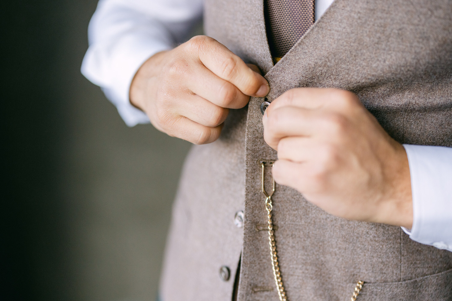 A close-up of hands buttoning a brown vest with a chain accessory, showcasing fine tailoring and attention to detail.