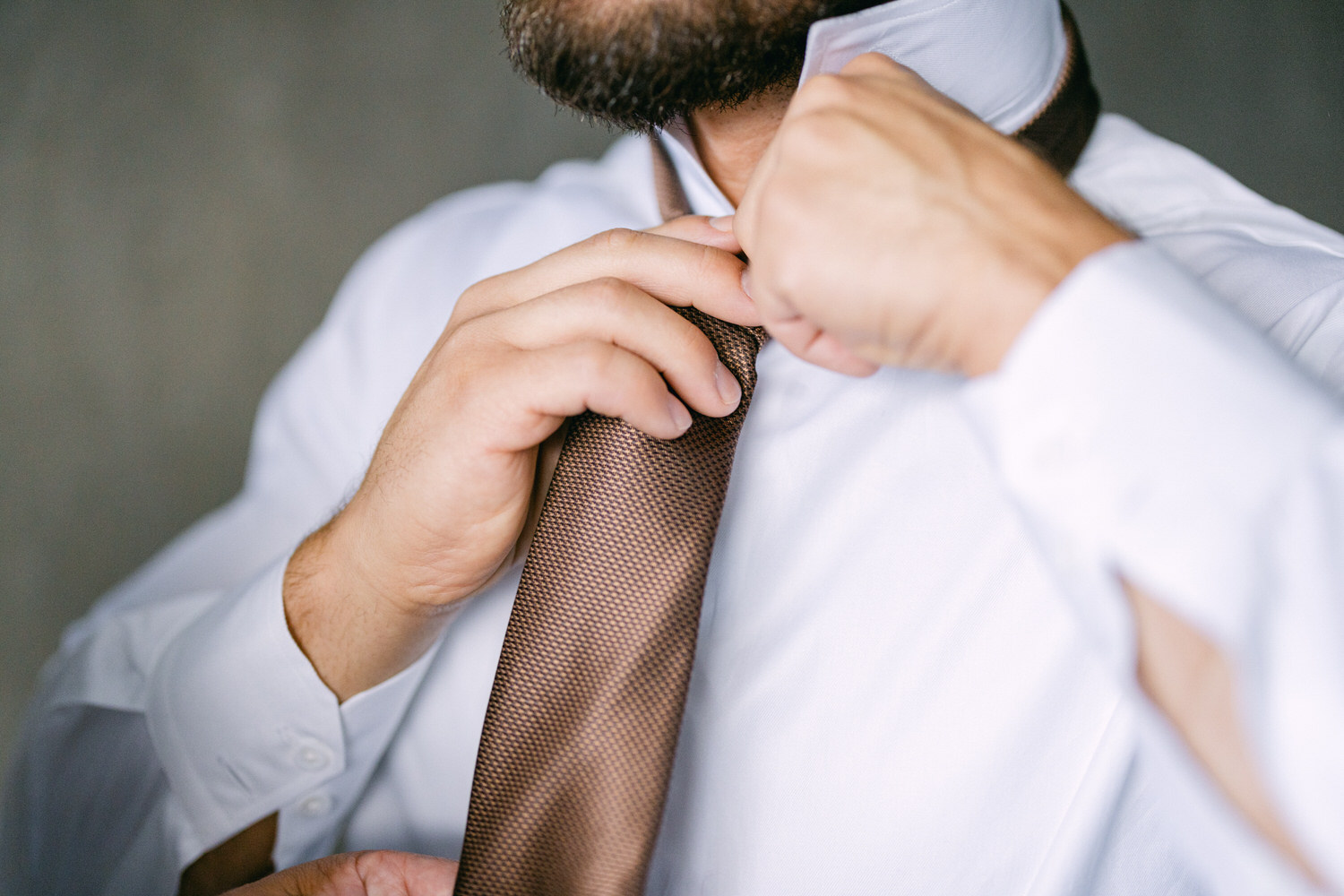 A close-up of a man adjusting a brown tie while wearing a white dress shirt, focusing on his hands and tie.