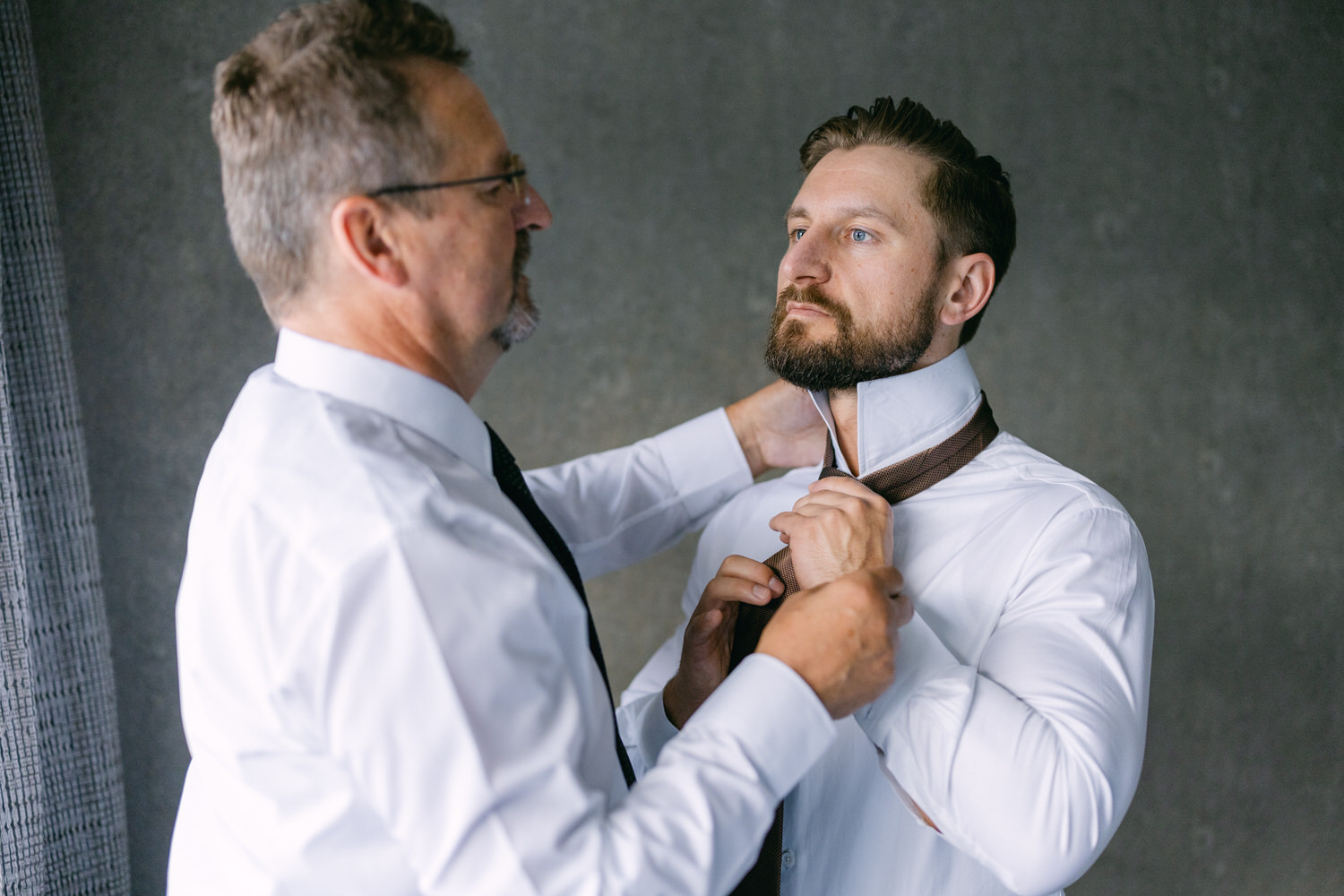 A man has his tie adjusted by another man, both wearing dress shirts in a modern setting.