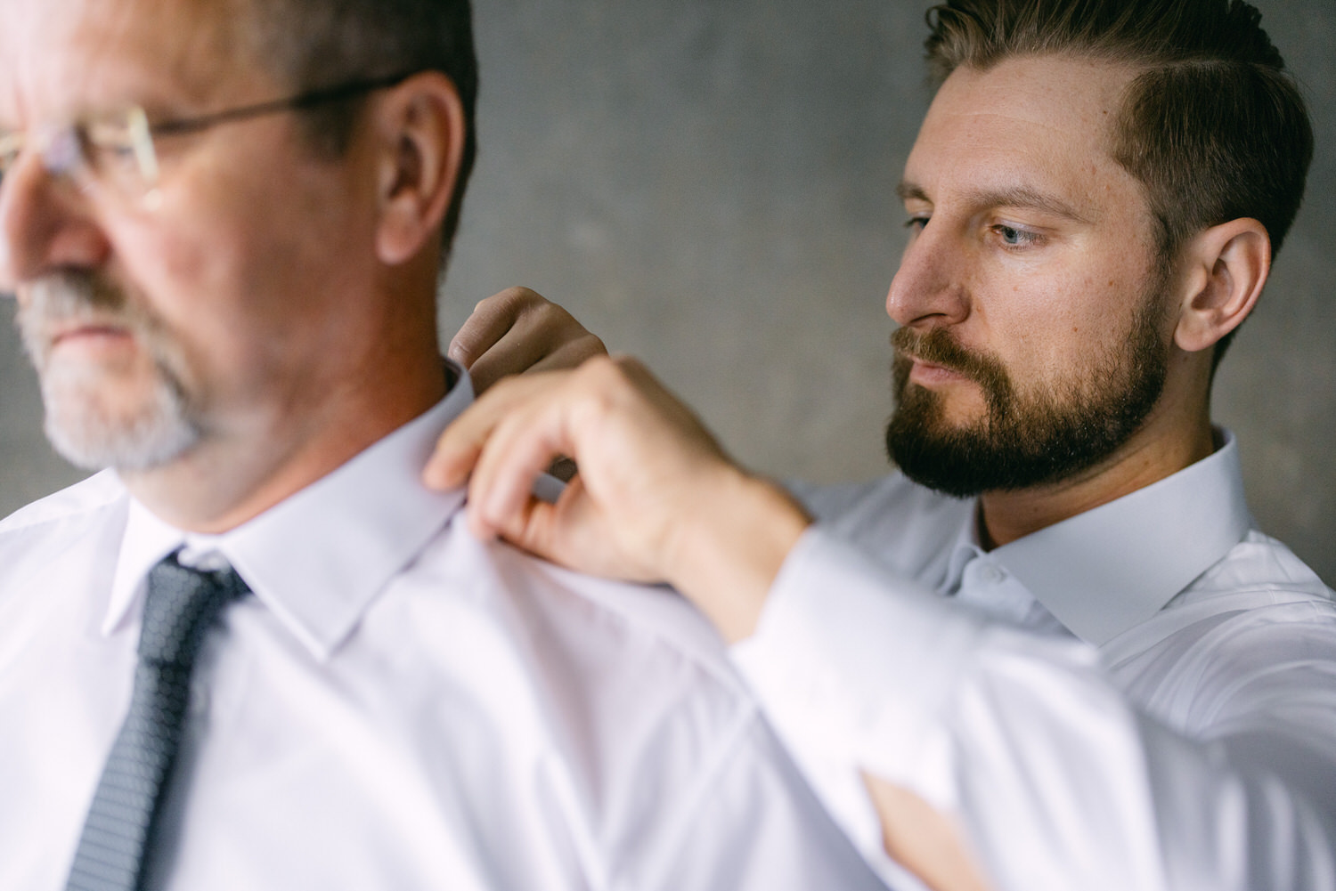 A young man adjusts the collar of a shirt for an older man, showcasing a moment of preparation and attention to detail.