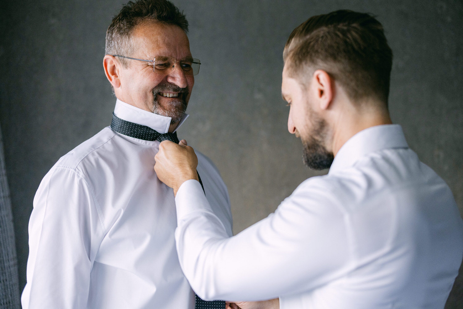 A young man helps an older man adjust his tie as they prepare for an event, both wearing white shirts in a stylish setting.