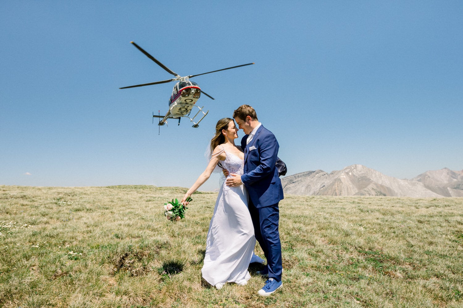 A bride and groom share a romantic moment outdoors while a helicopter flies overhead, with mountains in the background.