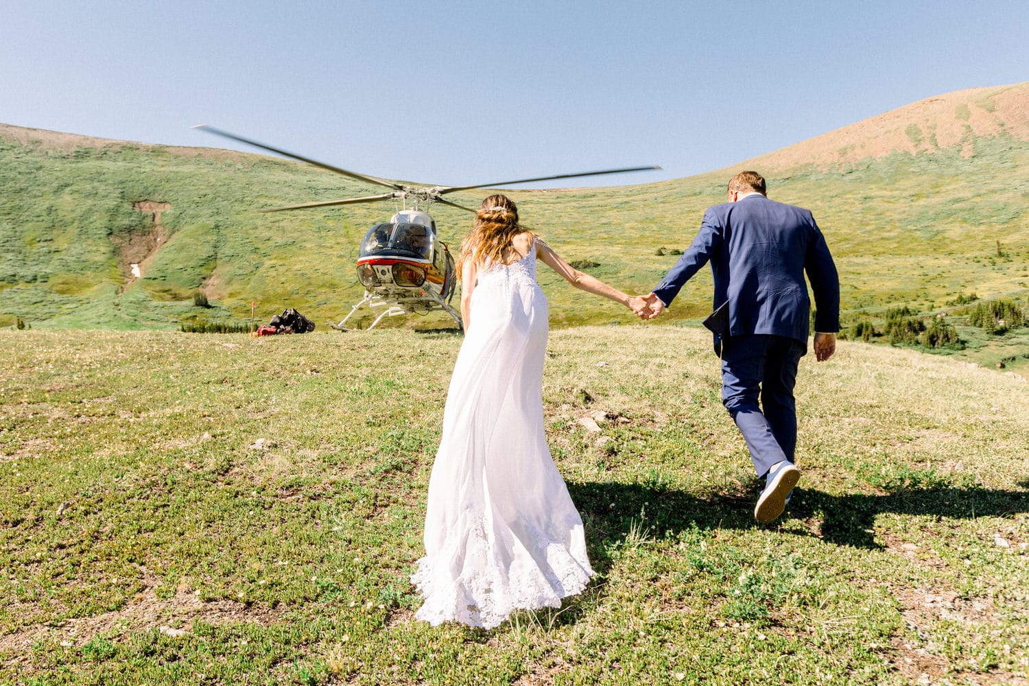 A couple in wedding attire holding hands while walking towards a helicopter on a grassy hillside.