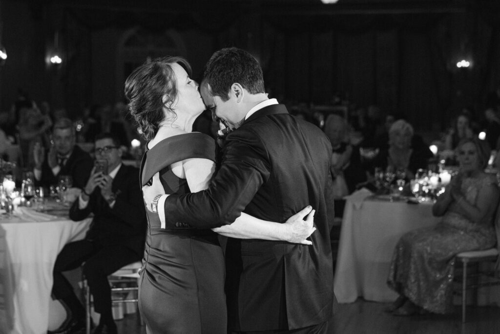 A woman kisses a man's forehead as they share a dance, surrounded by an audience of applauding guests in a dimly lit ballroom.