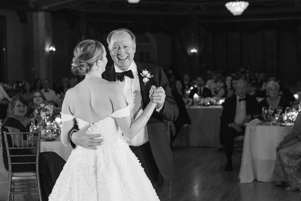 A joyful moment captured during a father-daughter dance at a wedding reception, with guests enjoying the celebration in the background.