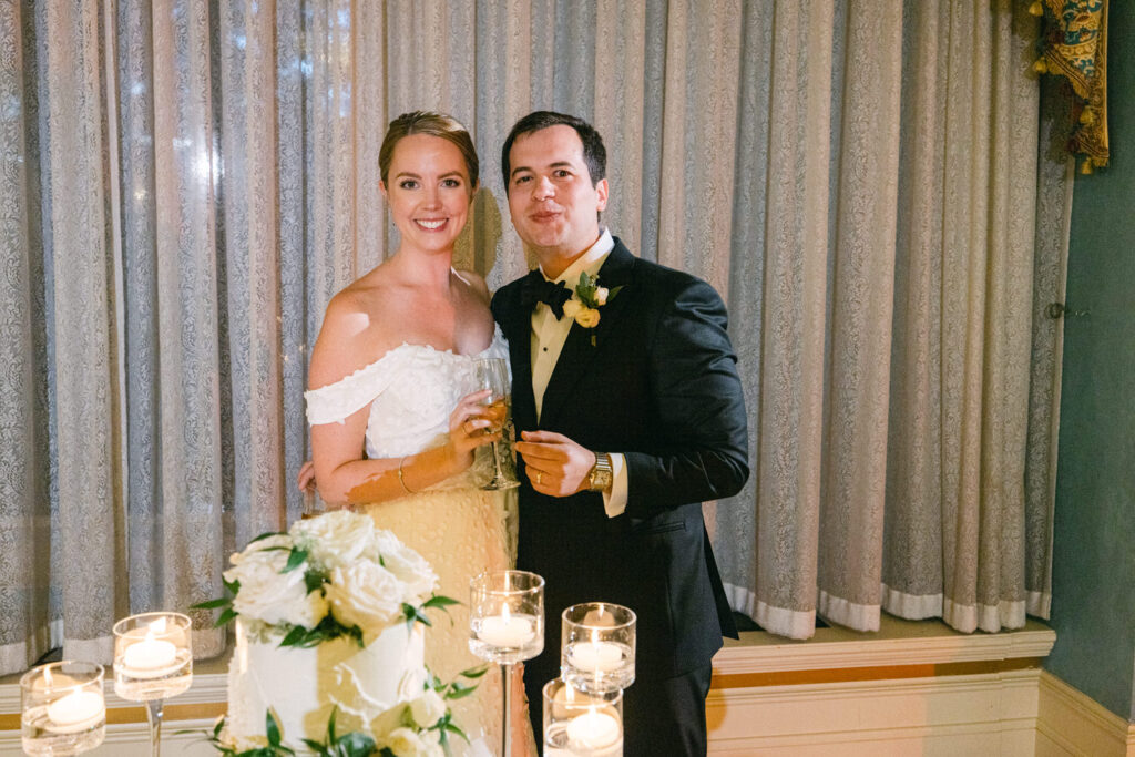 A happy couple stands together, smiling at the camera, with a wedding cake and candles in the foreground. The woman is wearing a white off-shoulder wedding dress, and the man is dressed in a classic black tuxedo.