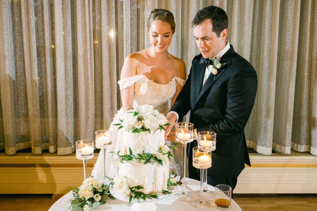 A bride and groom joyfully cutting a beautifully decorated wedding cake, surrounded by candles and floral arrangements.