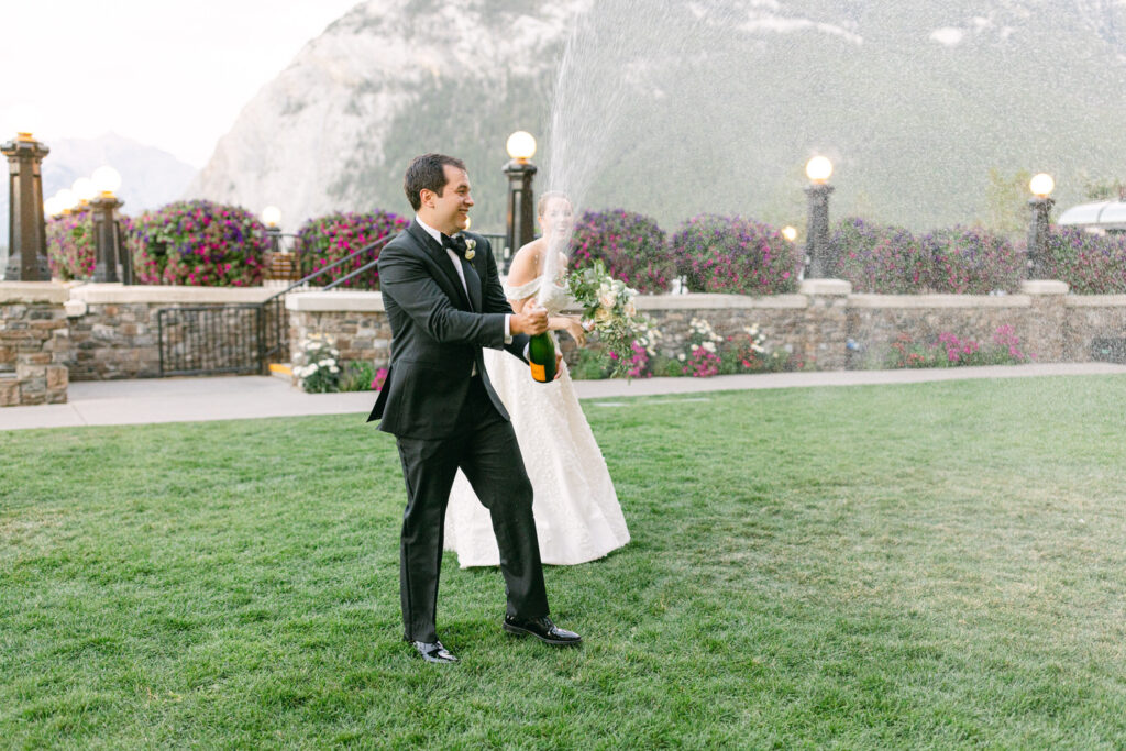 Celebratory Champagne Toast::A couple joyfully spraying champagne outdoors, surrounded by lush greenery and floral decorations, capturing a moment of celebration.