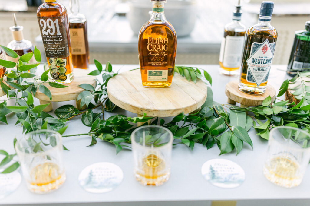 A collection of premium whiskey bottles displayed on wooden platforms, surrounded by fresh greenery and empty glasses.