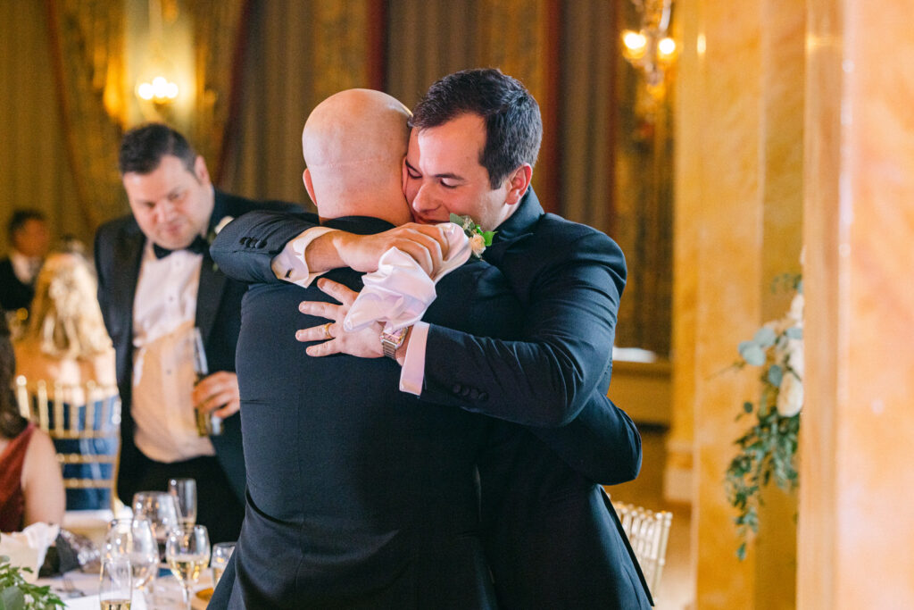 Two men embracing tightly at a formal event, capturing a heartfelt moment amidst guests and elegantly set tables.