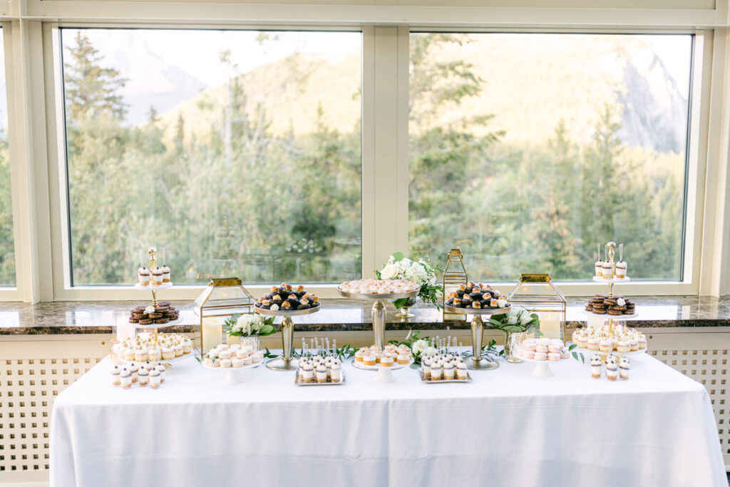 A beautifully arranged dessert table featuring an assortment of cupcakes, macarons, and pastries, set against a backdrop of mountains and greenery through large windows.