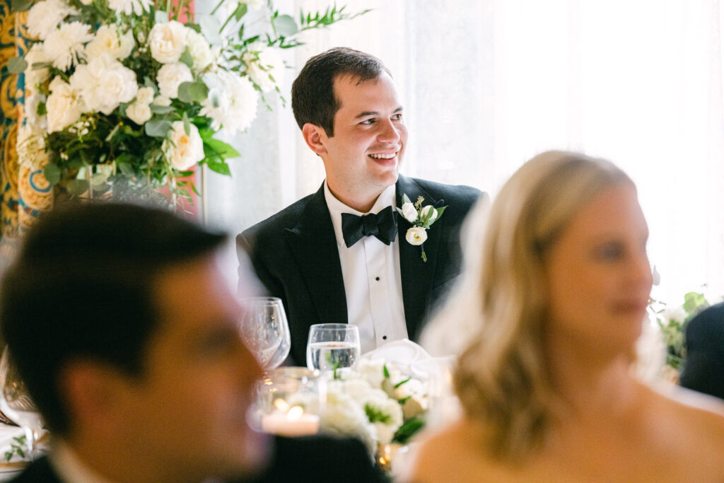 A smiling man in a tuxedo enjoys a cheerful moment at a formal event, surrounded by guests and elegant floral arrangements.