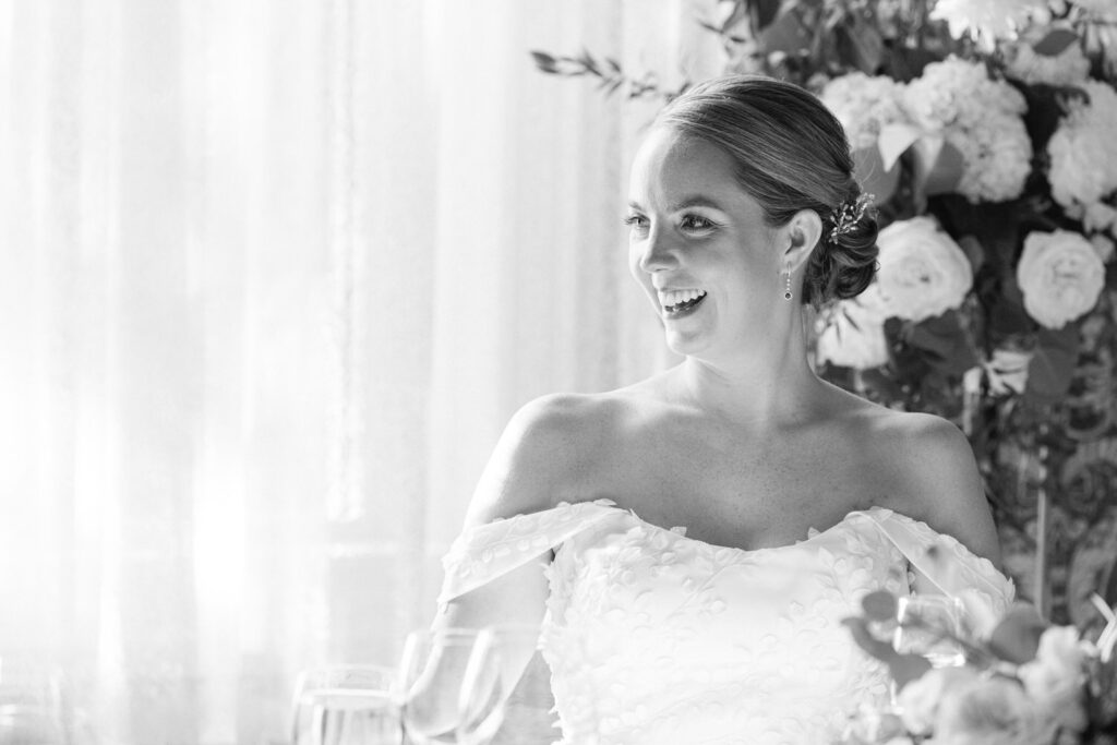 A smiling bride wearing an elegant off-the-shoulder dress, surrounded by floral arrangements and soft natural light.