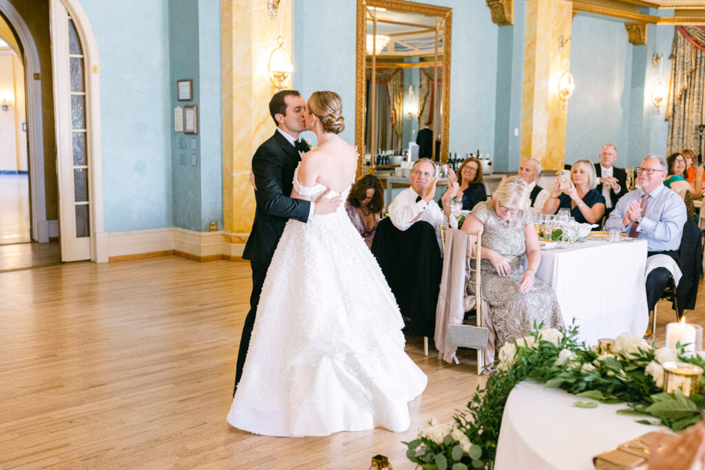 A couple shares a romantic kiss during their wedding reception, surrounded by applauding guests in an elegantly decorated venue.