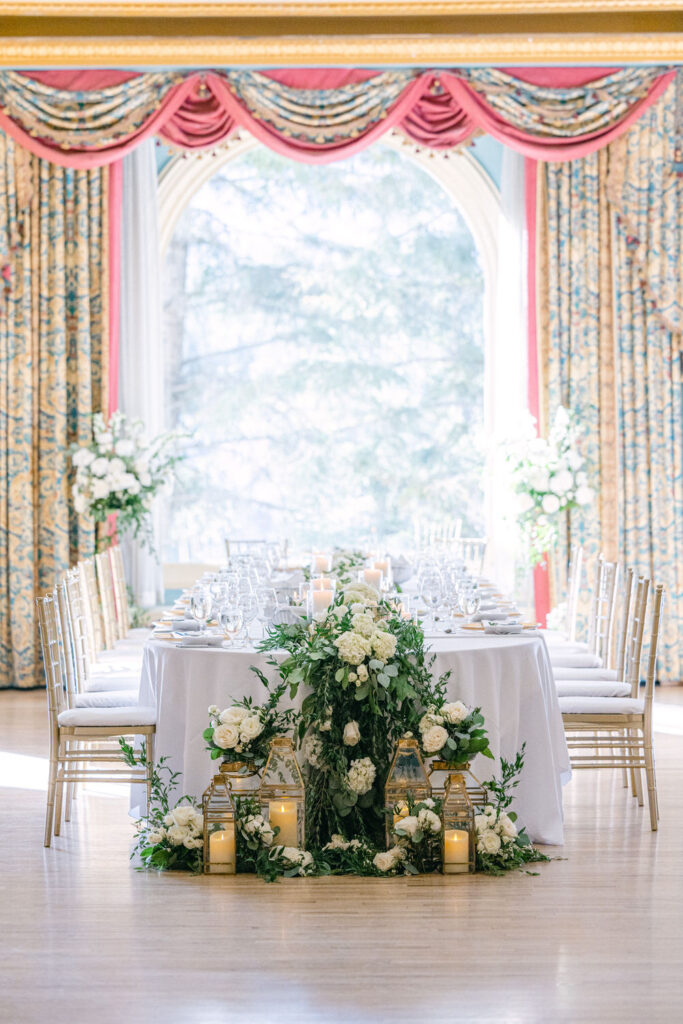 A beautifully arranged dining table adorned with floral decorations, candle lanterns, and an elegant setting in a well-lit room with patterned curtains.