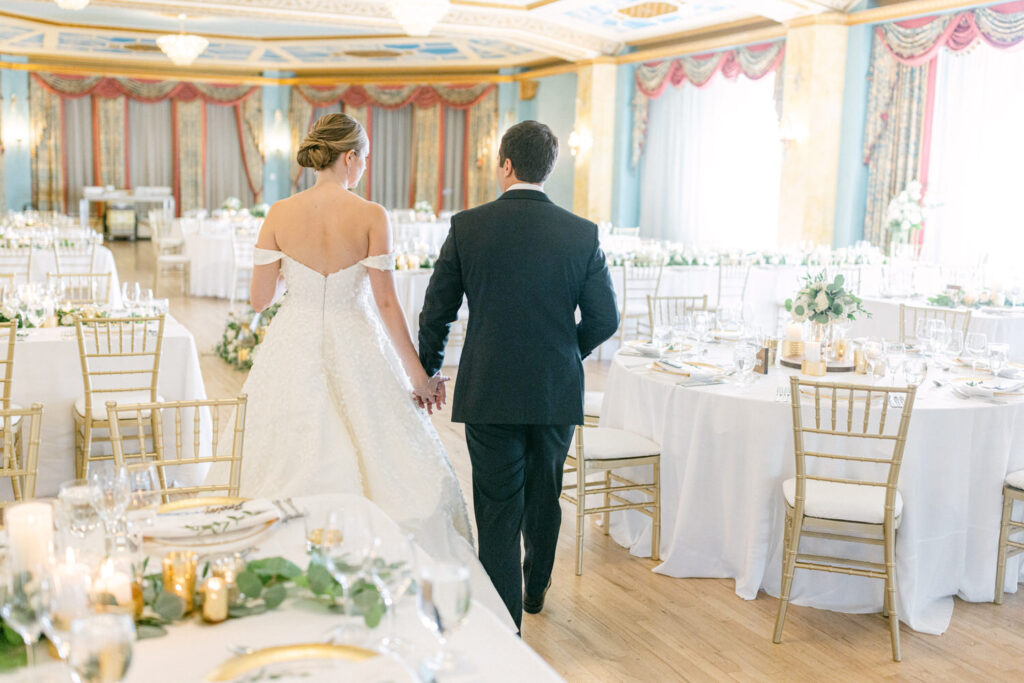 A bride and groom walking hand-in-hand through a beautifully decorated reception hall, featuring elegantly set tables and soft lighting.