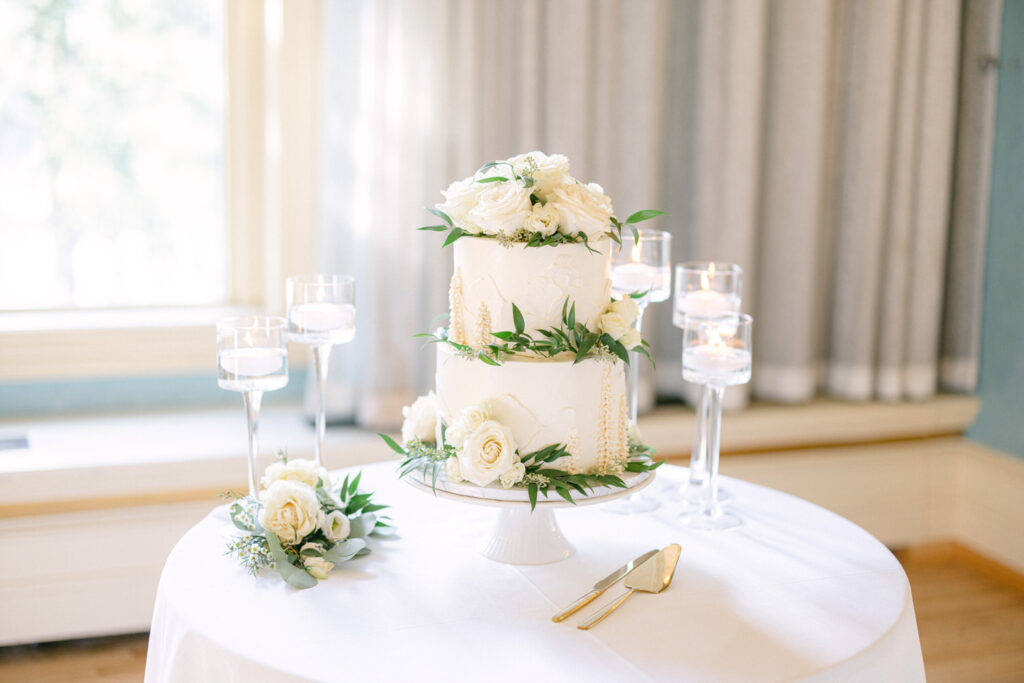 A beautifully decorated three-tier wedding cake adorned with white roses and greenery, surrounded by elegant candle holders on a white tablecloth.